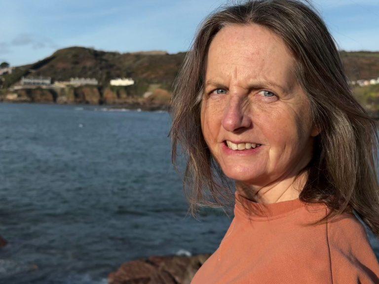 Jayne is leaning on a signpost which is pointing to the South West Coast Path. She's wearing a yellow hoody with the motif "Take it easy"